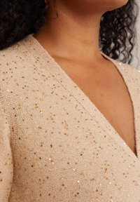 Bež pleten pulover z globokim V-izrezom, s razpršenimi zlatimi bleščicami, mehkobo in prilagojenim silhueto.