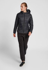 Czarna kurtka puchowa z przodu na zamek i kapturem, z pikowanymi detalami. W zestawie z czarnymi spodniami i czarnymi sneakersami, pokazana w ruchu.