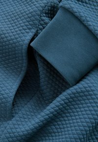 Tessuto teal texture con un motivo a diamante; include un dettaglio a polsino liscio e monocolore. Aspetto morbido e trapuntato, adatto per abbigliamento.