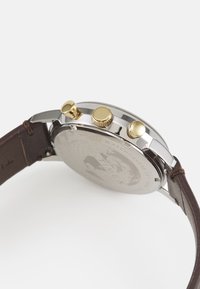 Montre en métal argenté avec trois poussoirs dorés, bracelet en cuir marron et arrière texturé présentant un motif gravé.