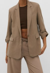 Femme portant un blazer beige avec manches retroussées, un pantalon à cordon assorti, et un haut court marron foncé, dévoilant un piercing au ventre.
