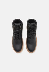 Botas de tobillo de cuero negro con superficie texturizada, puntera redonda, ojales de metal y suelas de goma marrón. Los cordones pasan a través de los ojales.