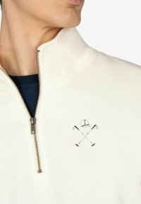 Giacca a zip in pile bianco con colletto alto e zip dorata. Dettaglioembroidero di mazze incrociate e berretto sul davanti.