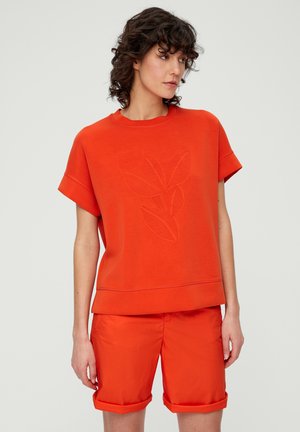 Oranje sweatshirt met korte mouwen met een bladpatroon; gecombineerd met bijpassende oranje shorts die om de mouwen zijn opgerold. Soepele textuur, casual ontwerp.