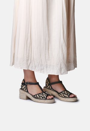 Stopy w czarnych, plecionych sandałach na koturnie z paskiem w kształcie litery T i beżowymi podeszwami z juty, zestawione z długą, zwiewną, jasną spódnicą.