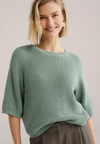 Pull en maille vert clair avec manches courtes et amples, texture côtelée, et encolure ronde. Associé à un pantalon foncé et fluide.