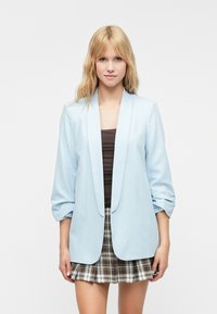 Svetlomodrý blazer so šálovým golierom a vyhrnutými rukávmi, kombinovaný s hnedým topom a plisovanou kockovanou mini sukňou.