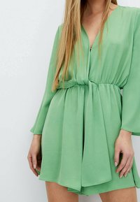 Vestido verde, ligero, con mangas largas, escote en V profundo, cintura elástica y detalle atado en la parte frontal. Textura suave y diseño fluido.