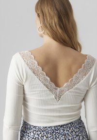Hvitt pliisert langermet topp med en dyp V-hals i blonder. Blonden har blomsterdesign. Bakgrunn i ensfarget grått, ingen ytterligere detaljer synlige.