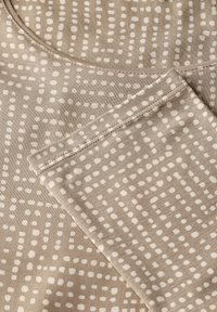 Beige Stoff mit einem gepunkteten Muster, das abwechselnd kleine und große Punkte zeigt. Glatte Textur, rundhals und gestickte Nähte.