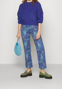 Stickad lila tröja, blå mönstrade byxor med abstrakta motiv, gröna klumpiga loafers och en vävd blå handväska.