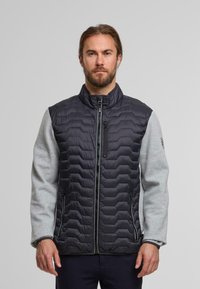 Veste rembourrée avec un corps en tissu matelassé noir et des manches en polaire grise. Elle possède un col montant, une fermeture éclair et des poches latérales.