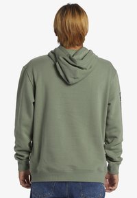 Felpa verde oliva con finitura texturizzata, grande cappuccio e polsini a costine. Presenta un logo stampato sulla manica destra. Vestibilità casual.