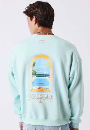 Mann trägt hellblauen Sweatshirt mit einer Grafik von einem Pool und Meerblick, eingerahmt von einem Fenster, mit Text "Luxury Life Dreaming of Monaco."