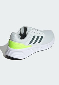 Chaussure de sport blanche avec un upper en maille texturée, un accent vert près du talon et une semelle en caoutchouc noire ornée de trois bandes grises sur le côté.