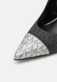 Chaussure noire pointue avec embout argenté texturé présentant un motif géométrique répétitif, shown against un fond blanc.