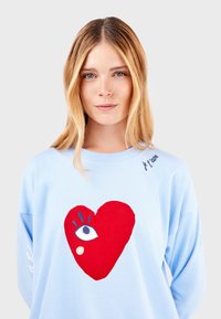 Sudadera azul claro con un gran gráfico de un corazón rojo con un ojo estilizado y la frase "je t'aime" en el hombro. Textura suave.