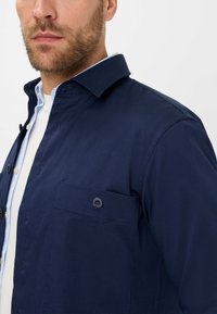 Chemise bleu marine à boutons avec un col pointu, une poche poitrine à deux boutons, et une texture lisse. Elle présente un col intérieur contrastant en bleu clair.