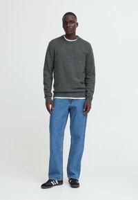 Grauer gestrickter Pullover mit Rundhalsausschnitt, über einem weißen Hemd getragen, kombiniert mit blauen Jeans und schwarzen Sneakers mit weißen Akzenten.