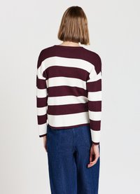 Person mit kurzen braunen Haaren, der einen langärmligen, rot-weißen gestreiften Pullover und blaue Jeans trägt, mit dem Rücken zu einem schlichten Hintergrund gewandt.