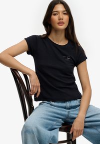 Mujer joven con cabello largo y oscuro, vestida con una camiseta negra y jeans azul claro, sentada de manera relajada en una silla de madera sobre un fondo blanco.