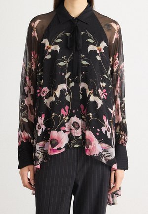 Schwarze transparente Bluse mit pinken und weißen Blumen- und Vogelmustern, langen Ärmeln, Schleifenkragen, getragen zu dunklen Nadelstreifenhosen.