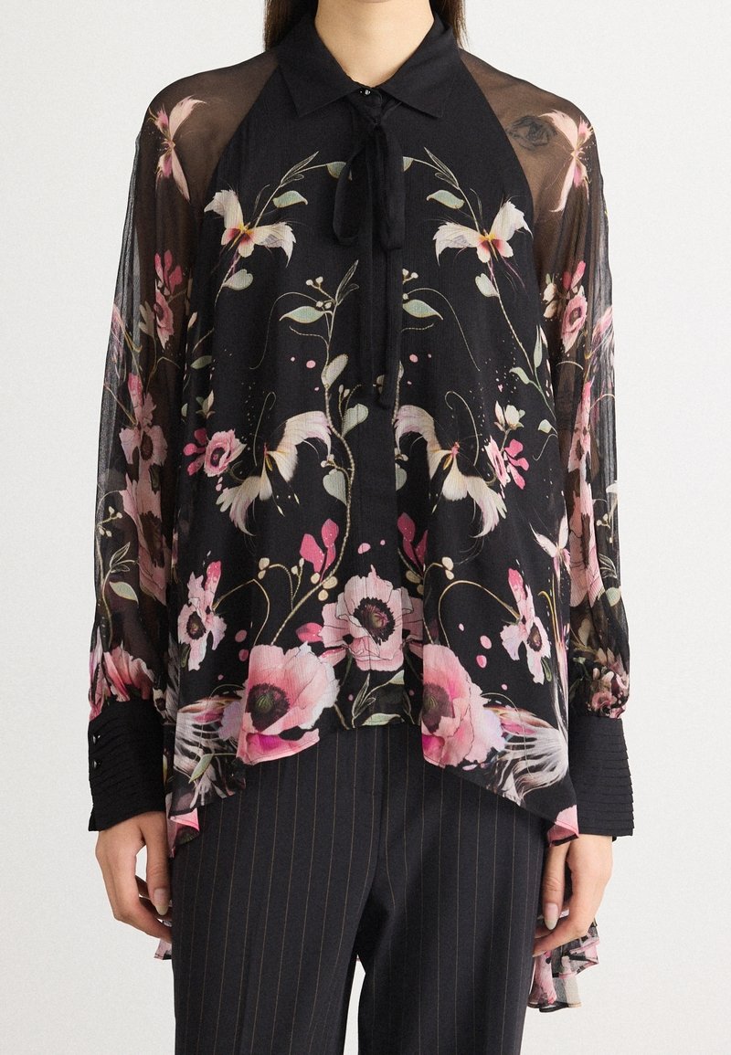 Blouse noire transparente avec motifs floraux et d’oiseaux roses et blancs, manches longues, col à nouer, portée avec un pantalon sombre à fines rayures.