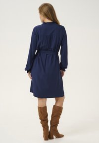 Robe bleu marine avec une taille ceinturée, manches longues avec des poignets froncés et un détail froncé dans le dos, associée à des bottes marron à hauteur de genou.