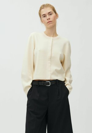 Donna con le mani nelle tasche che indossa un maglione crema con bottoni e pantaloni neri con cintura nera su uno sfondo semplice.