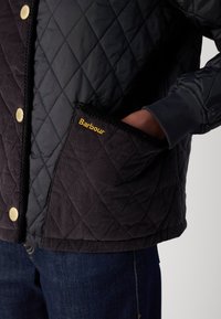 Quilted black jacket with gold hardware and a corduroy pocket featuring "Barbour" lettering. The fabric has a smooth, matte texture.