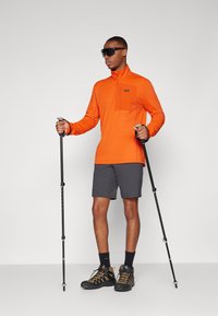 Spožs oranžs garām piedurknēm top ar rāvējslēdzēja apkakli, pelēkas šorti, melni kalnu apavi un trekinga nūjas. Mūsdienīgs sporta dizains, teksturēta auduma.