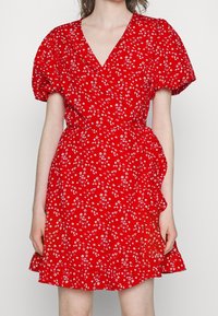 Mujer vistiendo un vestido envolvente rojo con mangas abullonadas y estampado floral blanco, de pie contra un fondo blanco liso.