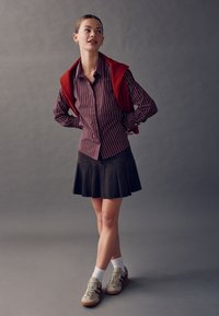 Bordó csíkos, gombos ing, piros pulóver átvetve, fekete pliszírozott szoknya, fehér bokazokni és bézs sportcipő bordó részletekkel.