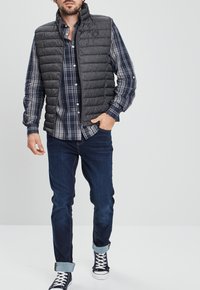 Chaleco de quilt gris sobre una camisa a cuadros azul y blanca, combinado con jeans oscuros y zapatillas navy con suelas blancas.