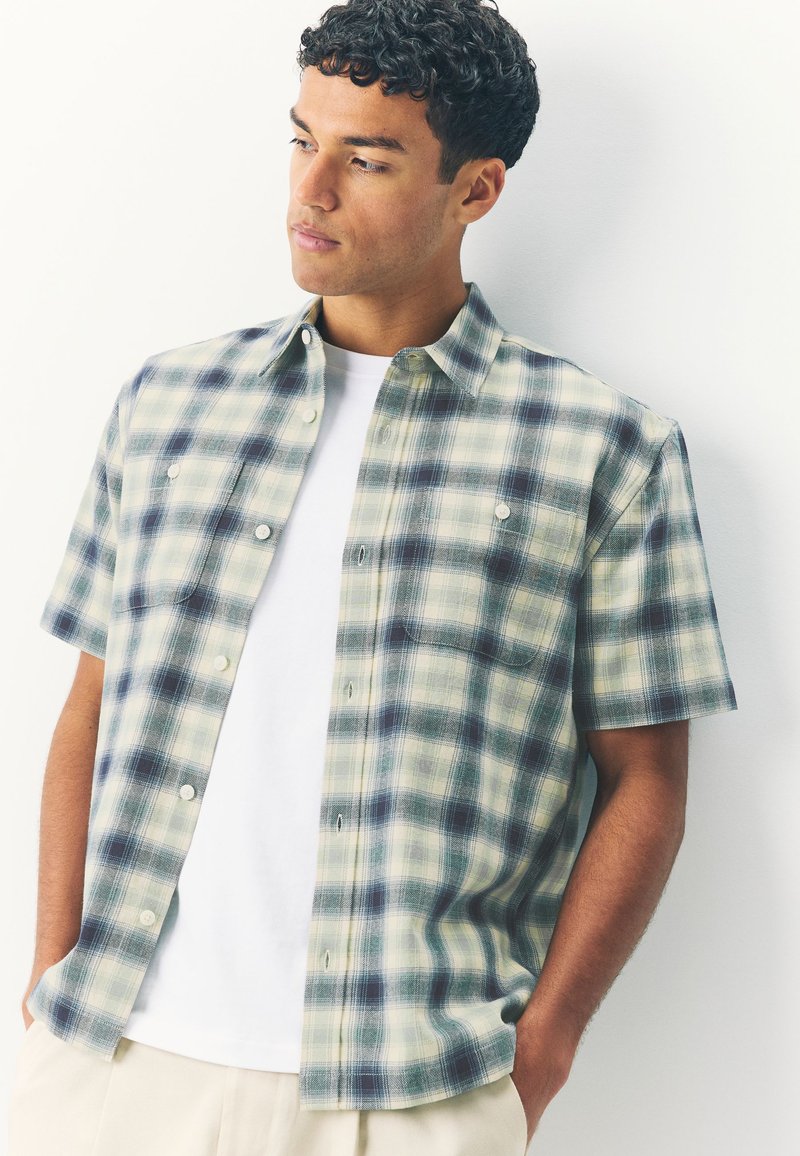 Short-sleeve plaid shirt in green and navy tones, featuring a button-up design and two chest pockets, worn over a white t-shirt.