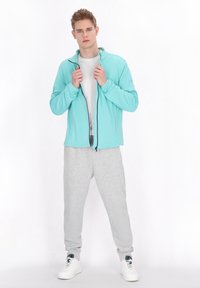 Giacca zip-up azzurro chiaro con colletto alto, abbinata a pantaloni sportivi grigi. Il modello si sta posizionando con le mani sulla giacca, indossando sneakers bianche.