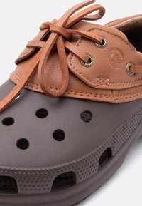 Καφέ και γκρι λαστιχένιο σανδάλι με οπές αερισμού, δερμάτινα κορδόνια σε ταμπέ απόχρωση δεμένα σε φιόγκο και χάλκινα περικάρπια με λεπτομέρειες επωνυμίας.