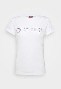 Camiseta de algodón blanca con letras plateadas reflectantes en negrita que dicen "HUGO" en el frente. Diseño de mangas cortas y cuello redondo.