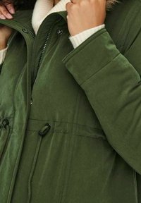 Parka verte à texture douce, équipée d'une fermeture éclair et de boutons-pression, taille avec cordon de serrage et détail de capuche doublée de fourrure.