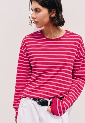 Jeune personne aux cheveux courts et foncés portant une chemise rayée rose et rose clair à manches longues et un pantalon blanc avec une ceinture noire.