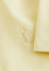 Tejido texturizado amarillo pálido con un sutil detalle bordado de cabeza de ciervo y un borde doblado de una prenda.
