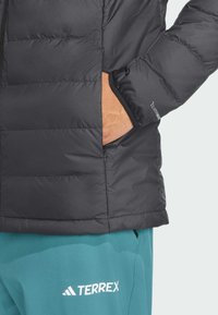 Black puffer jacket with quilted texture and a zippered pocket. Features adjustable cuffs with a logo on the sleeve. Paired with teal pants.