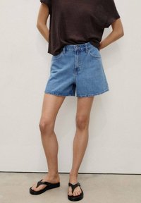 Personne portant un short en denim bleu taille mi, un haut sombre à manches courtes et des tongs noires, debout devant un mur clair uni.