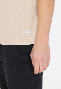 Beige t-shirt med en rund halsudskæring og et lille hvidt mærke på siden; båret med sorte bukser, hvor en hånd hviler på stoffet.