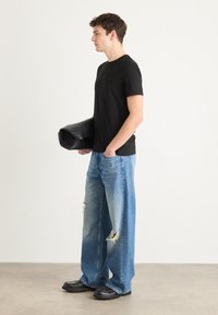 Zwarte T-shirt met korte mouwen, versleten wijde spijkerbroek in blauw, stevige zwarte schoenen, houdt een zwarte tas vast, staat tegen een neutrale achtergrond.