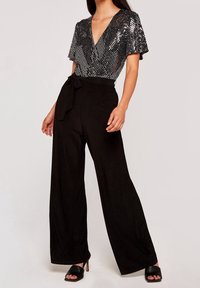 Pantalons noirs ample avec une texture satinée et une ceinture à nouer, associés à un haut scintillant en sequins argentés avec un décolleté en V profond.