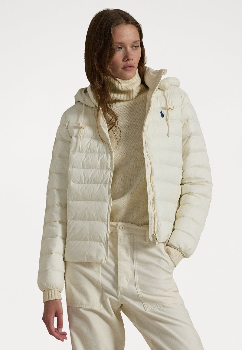 Puffer jacket in cream, featuring a zippered front, adjustable drawstring hood, and rib-knit cuffs, worn with a beige turtleneck and pants.