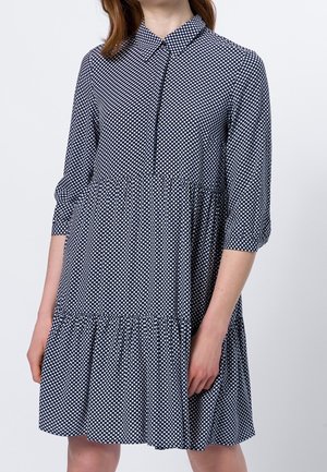 Vrouw draagt een marineblauwe jurk met witte ruitpatroon, driekwart mouwen, een kraag en een geplooide, gelaagde rok.