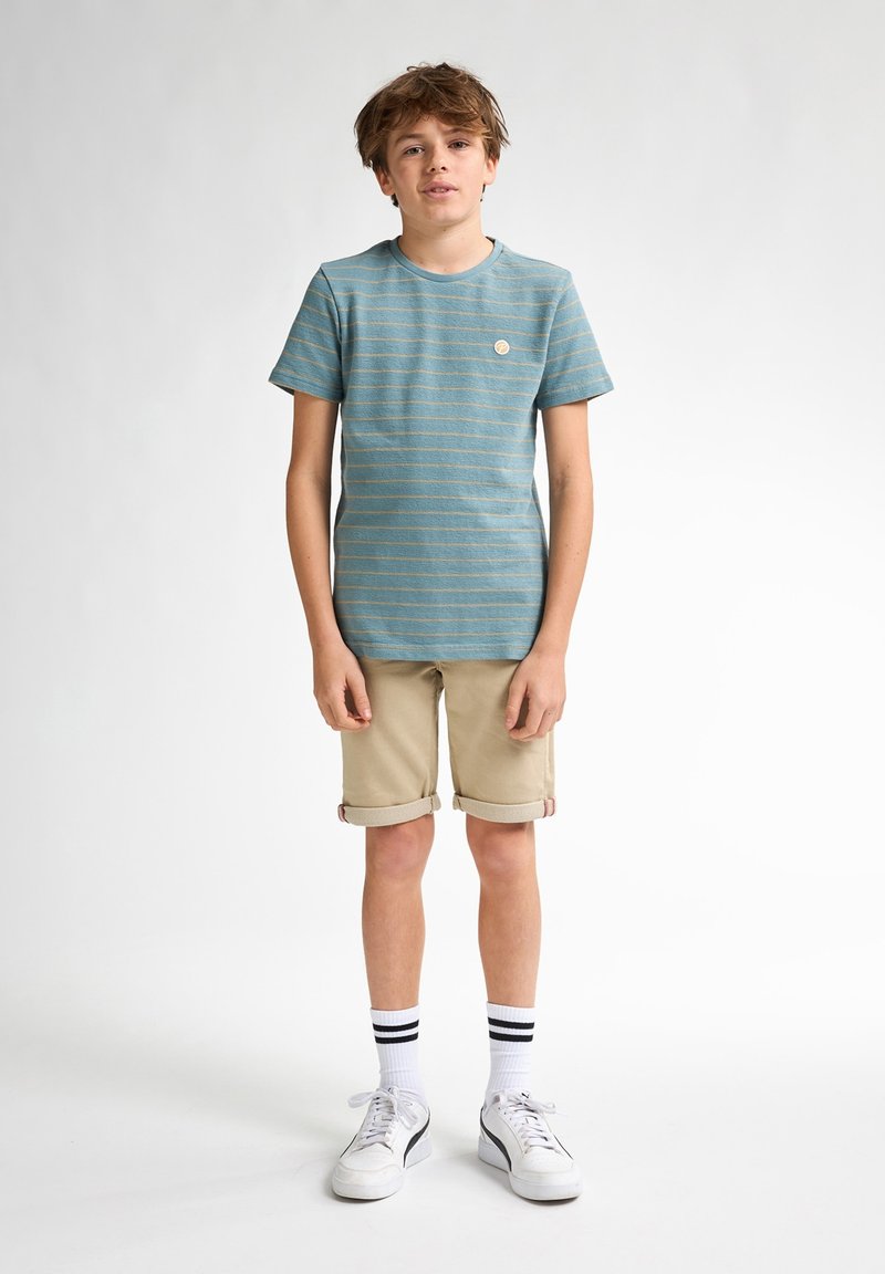 Camiseta de manga corta a rayas en color teal con un pequeño logo circular, combinada con pantalones cortos beiges enrollados y zapatillas blancas con rayas negras.