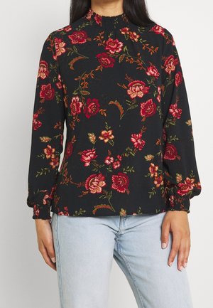 Frau trägt eine schwarze Langarmbluse mit roten und pinken Blumenmustern und hellblauen Jeans, steht vor einem schlichten Hintergrund.